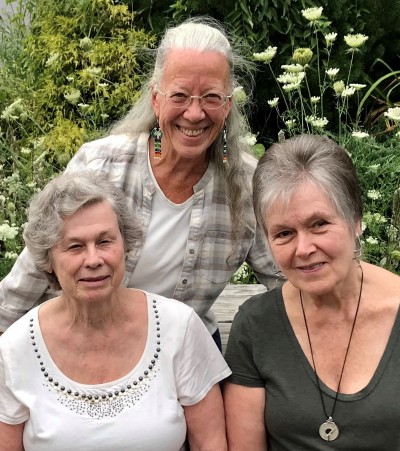 This is a photo of three three women who founded the Holistic Health Community. Fay Loomis has short wavy silver hair and wears a white scoop-necked shirt. Dr. Nancy Eos has long white hair, glasses and long earrings. Cornelia Wathen has short salt-and-pepper hair and is wearing a pendant and a slate-gray shirt. All three women are smiling.
